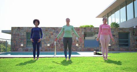Three women practicing outdoor yoga on sunlit lawn by modern pool for wellness
