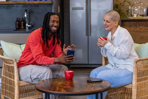 Multicultural friends laughing while sharing smartphone in cozy kitchen with red mugs