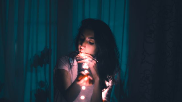 Woman holding and admiring fairy lights in a dark, dimly lit room. Warm light from the fairy lights contrasts with the surrounding darkness, creating a soft, dreamy ambiance. The curtains and background add to the relaxation and contemplative mood. Ideal for themes of tranquility, creativity, intimate home settings, and artistic expression.