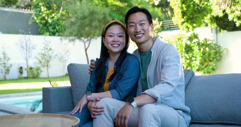 Asian couple relaxing on outdoor sofa by pool displaying matching smartwatches