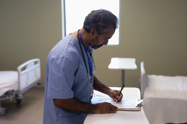 Biracial male doctor in a hospital ward writing on a clipboard. Ideal for healthcare, medical, and hospital-related content. Can be used in articles, brochures, and websites focusing on medical professionals, patient care, and hospital environments.