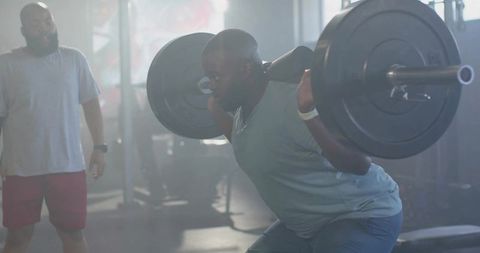 Male athlete performing heavy barbell back squat with spotter in functional gym