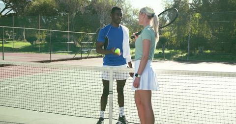 Diverse tennis players chatting at net on sunlit public court holding racket and ball