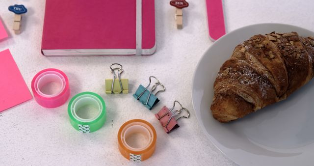 A delicious croissant paired with office supplies creates a scene of a casual work breakfast, with copy space. Various stationery items like sticky notes, clips, and a notebook suggest a productive setting with a touch of relaxation.