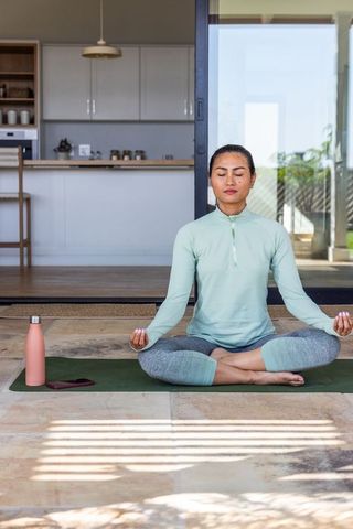 Asian woman meditating on sunlit patio in mint activewear practicing mindfulness and calm