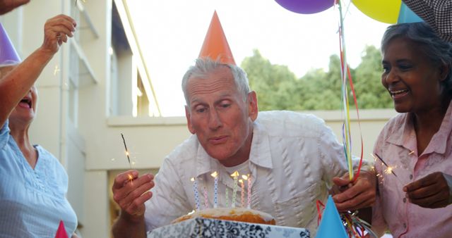 A middle-aged Caucasian man blows out candles on a birthday cake, surrounded by diverse friends celebrating, with copy space. Joyful expressions and party hats contribute to the festive atmosphere of the birthday gathering.