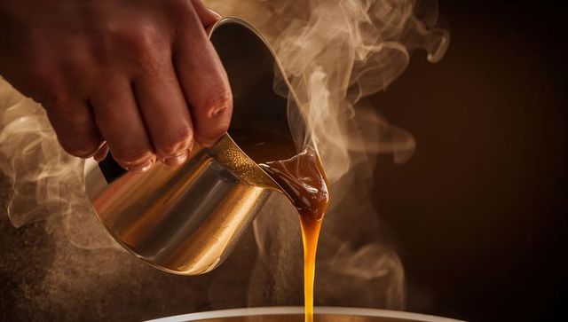 Pouring steaming espresso from stainless steel milk pitcher with rich caramel crema closeup