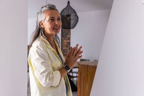 Middle-aged woman standing by doorway, clasping hands, wearing smartwatch and hoop earrings, bright