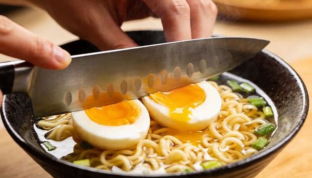 Hands slicing soft-boiled egg over steaming ramen bowl with runny yolk