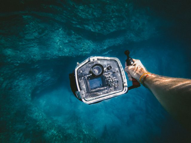 Person holding an underwater camera while exploring deep, clear blue ocean. Ideal for use in themes related to underwater exploration, scuba diving, marine life research, underwater photography, aquatic sports, and travel adventures.