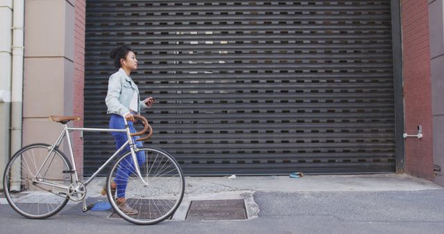 Woman walking her bicycle past a closed urban garage door. She wears a casual outfit, with a denim jacket and blue jeans. The scene captures city life and active lifestyle. Useful for content related to urban exploration, commuting, outdoor activities, or modern lifestyle.
