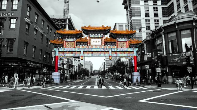 Depicting an intersection in a bustling urban Chinatown, this image features traditional and colorful gateway architecture standing amidst modern buildings. Pedestrians are crossing the street, adding to the dynamic city atmosphere. This image is ideal for topics involving urban culture, diversity, travel destinations, or photography featuring contrasts between traditional and modern architecture.