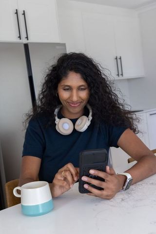 Smiling asian woman using smartphone at kitchen counter wearing headphones and smartwatch