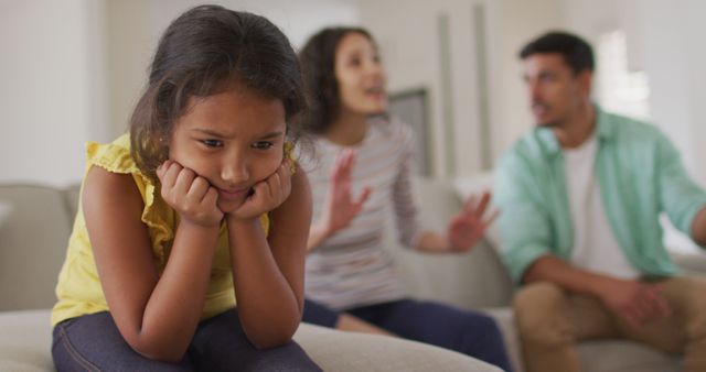 This stock photo depicts a young girl sitting on a couch with a forlorn expression while her parents are animatedly arguing in the background. She appears isolated and upset, highlighting the emotional impact of family conflicts on children. Suitable for use in articles or campaigns focused on family dynamics, childhood emotional well-being, and the effects of domestic disagreements on children.