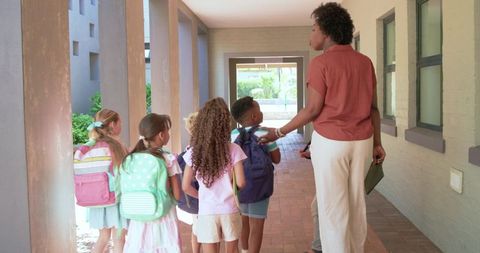 Female teacher guiding elementary students down school corridor with backpacks