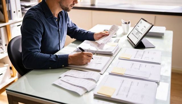 Man organizing invoices at glass desk with tablet and binder in bright home office