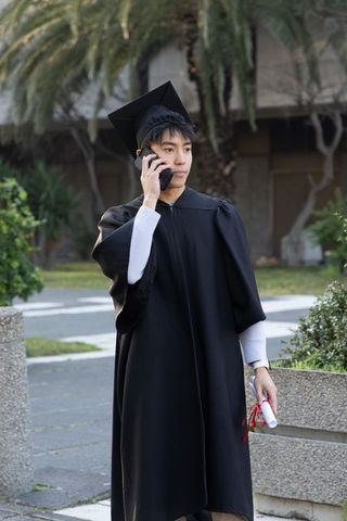 Asian graduate in mortarboard and gown holding diploma while making phone call on campus