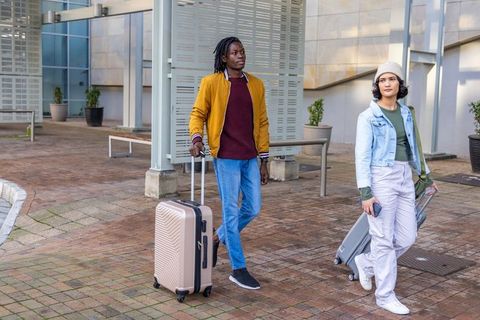 Young multicultural travelers walking urban plaza pulling suitcases and using smartphone