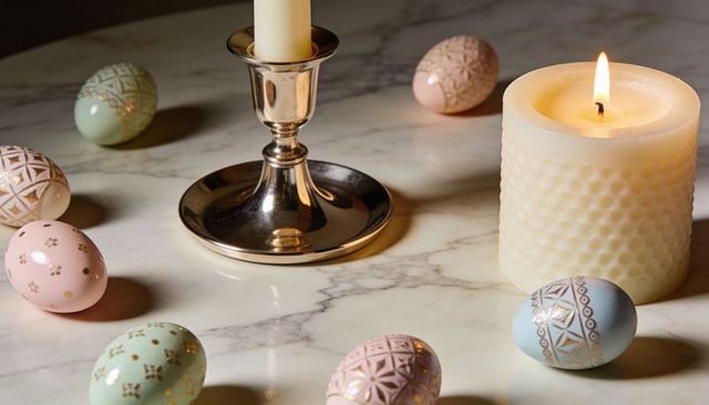 Burning cream candle glowing on marble with pastel decorated eggs and metallic candlestick