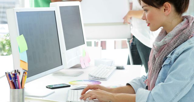 Woman concentrating on her work at a neatly organized desk with dual monitors. Ideal for depicting a modern office environment, productivity, creative professions, and workplace settings.