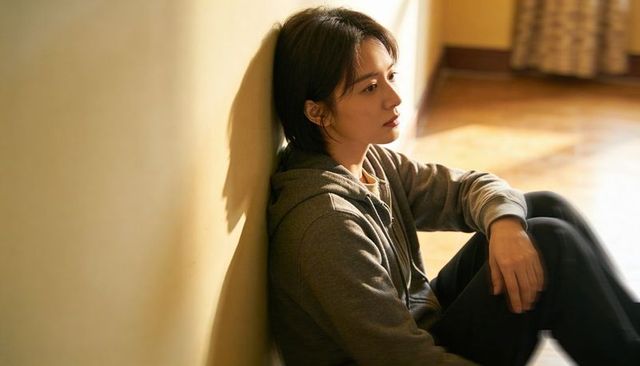 Contemplative young woman sitting against wall in sunlit room wearing gray hoodie