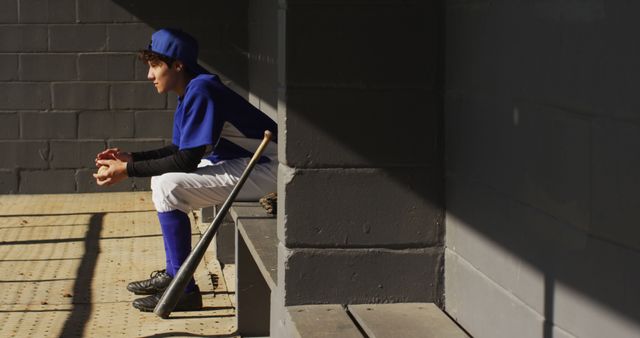 Young baseball player in uniform sitting in dugout, holding baseball, bat resting nearby. Useful for sports themes, youth sports programs, teamwork, and determination concepts.