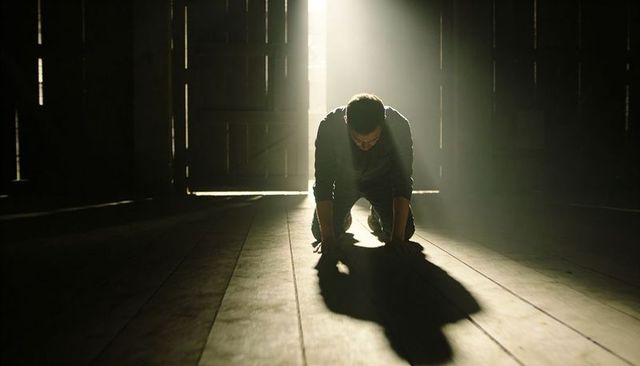 Man kneeling in dusty barn with sunbeam casting long shadow, moody cinematic light