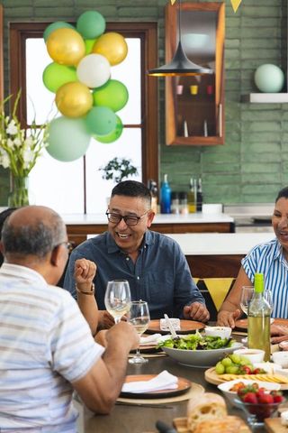 Senior asian family laughing and sharing meal at festive kitchen table with balloons and wine