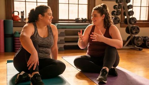 Two women chatting on yoga mats in sunlit gym, smiling and bonding after workout