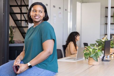 Confident black woman leaning on coworking desk with colleague working on laptop