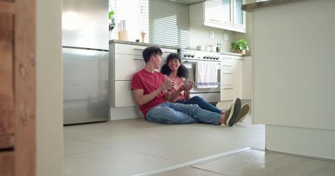 Diverse young couple sitting on kitchen floor holding mugs, enjoying cozy relaxed morning