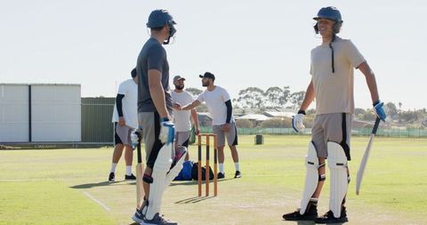 Diverse male teammates practicing batting with wooden bats and helmets on sunny cricket pitch