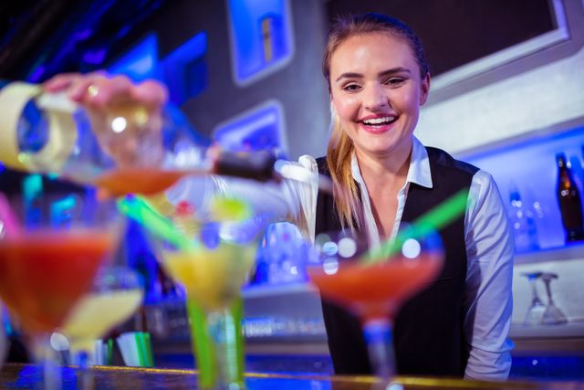A cheerful female bartender is seen pouring drinks into glasses at a bar counter. The vibrant lighting and colorful cocktails suggest a lively nightlife atmosphere. This image can be used for promoting bars, nightlife events, hospitality services, or articles related to mixology and bartending.