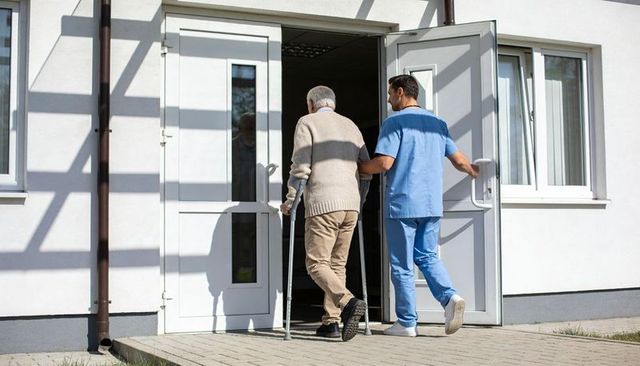 Caregiver guiding elderly man with forearm crutches entering medical clinic