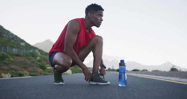 Athlete takes a moment to rest on an empty road against a picturesque mountain backdrop. Wearing casual sportswear, he remains hydrated with a water bottle placed next to him. This image is ideal for promoting sports brands, fitness goals, hydration products, or outdoor exercise routines showcasing determination and focus.