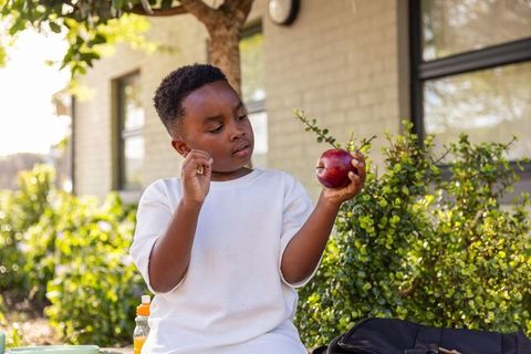 African american boy sitting on bench holding red apple at schoolyard in warm sunlight