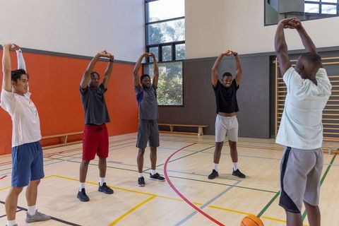 Diverse male teammates stretching arms overhead during basketball warm-up on indoor court