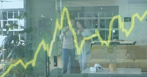 Two women reviewing tablet and analyzing yellow-green growth chart through office glass
