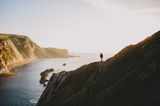 Person standing on cliff edge enjoying breathtaking coastal landscape at sunrise. Ideal for travel blogs, adventure trip promotions, inspiring nature photography collections, or eco-tourism advertisements conveying tranquility, natural beauty, and the spirit of exploration.
