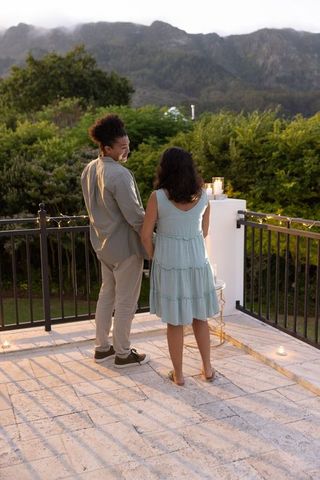 Diverse couple holding hands on rooftop terrace at dusk, gazing at mountain ridge