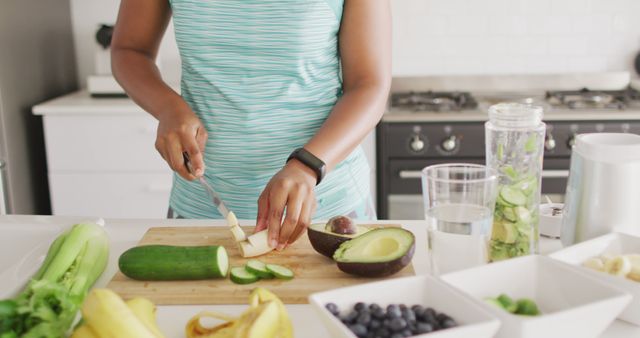 Woman in a modern kitchen preparing a healthy meal. She is slicing cucumber on a wooden board. Fresh ingredients like avocados, bananas, and blueberries are in bowls nearby. A blender with green smoothie ingredients is visible. Ideal for content related to healthy eating, recipes, wellness, and lifestyle blogs.