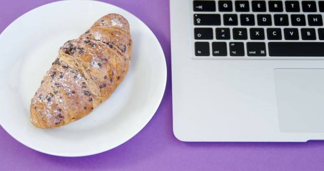 A chocolate chip croissant on a white plate sits next to a laptop on a purple surface, with copy space. It suggests a scenario of enjoying a snack or breakfast during a work break or while multitasking.