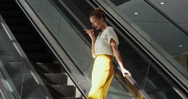 Business woman in white blouse and yellow skirt talking on phone while descending on escalator in modern building. Great for articles on professional communication, urban lifestyle, business environments, and modern work life.