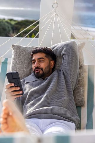 Indian man reclining on hammock holding smartphone on coastal balcony relaxing by ocean