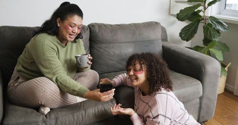 African american mom and daughter sharing smartphone on gray sofa wearing pink eye patches
