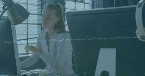 Woman analyzing data on dual monitors wearing headphones at bright office workstation