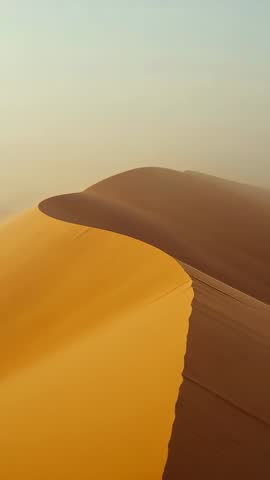 Gliding drone approaching sunlit desert dune crest with ripples and hazy distant ridges