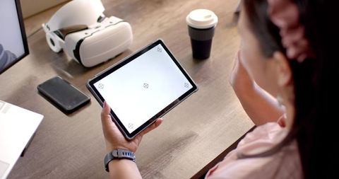 Woman holding tablet with blank screen at home office desk with vr headset, smartwatch, coffee
