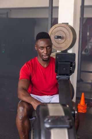 African american man rowing on ergometer in gym with weight plates and agility cone