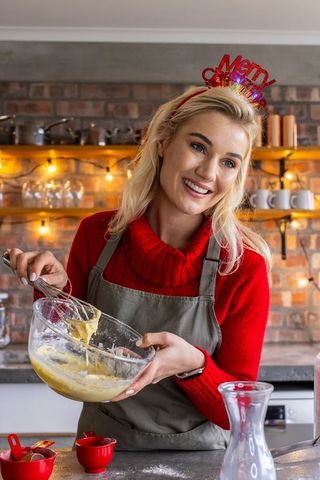 Smiling woman mixing holiday batter in cozy kitchen with brick wall and fairy lights